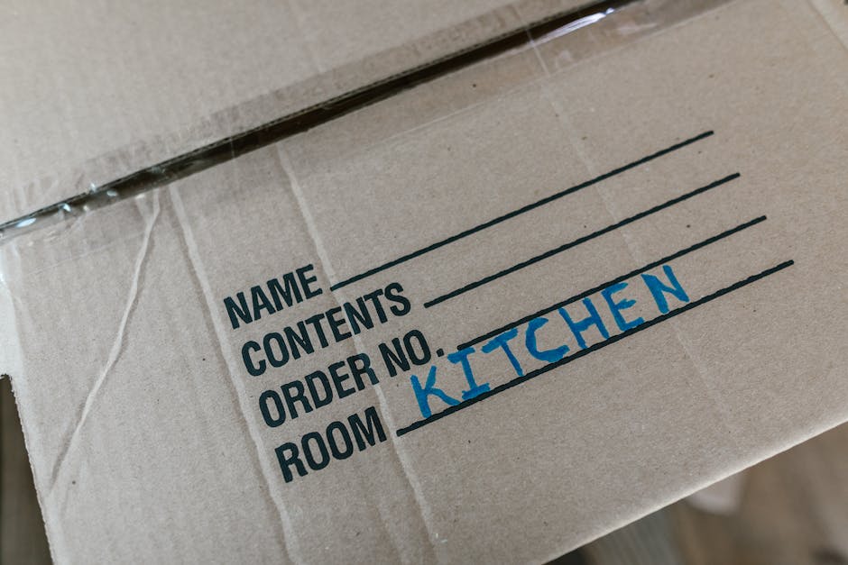 Close-up view of a brown cardboard moving box labeled with a handwritten blue marker that indicates the contents are from the 'kitchen'. The box is sealed with clear packing tape and features printed